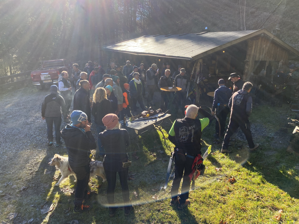 Gruppe von Bogenschützinnen und Bogenschützen vor Holzhütte im Sonnenschein